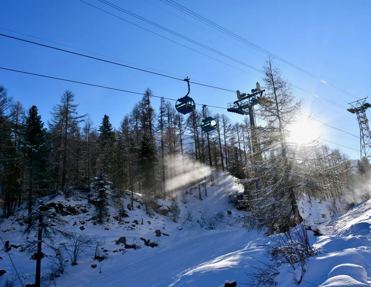 Teleférico con cabinas azules sobre una pista de esquí nevada con árboles y sol brillante.