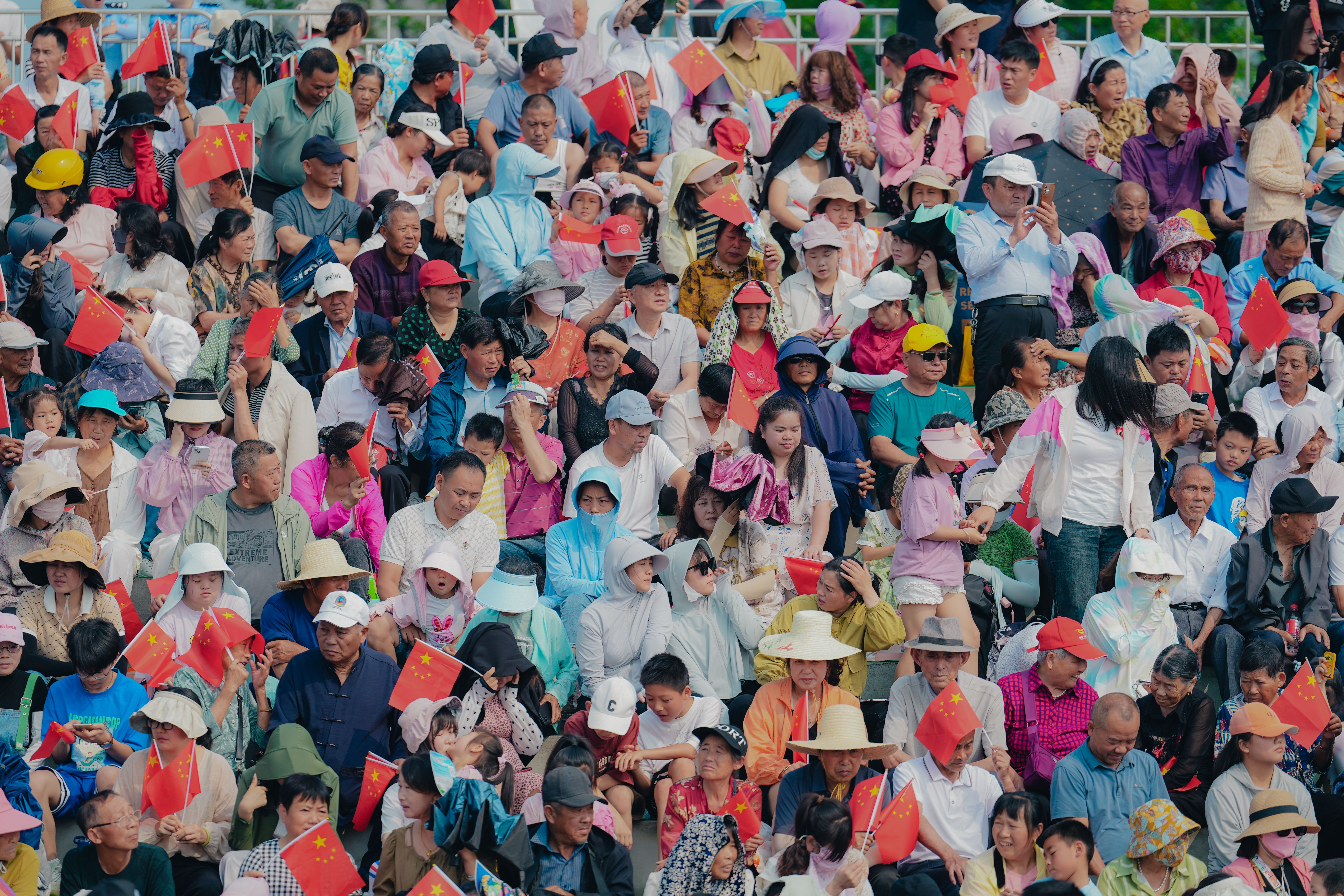 Multitud de personas de diversas edades sentadas en gradas, muchas sosteniendo pequeñas banderas chinas.