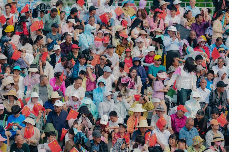 Multitud de personas de diversas edades sentadas en gradas, muchas sosteniendo pequeñas banderas chinas.