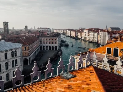 Vista panorámica de Venecia con edificios históricos de fachadas blancas y rojas junto a un canal, techos de tejas y campanarios lejanos bajo un