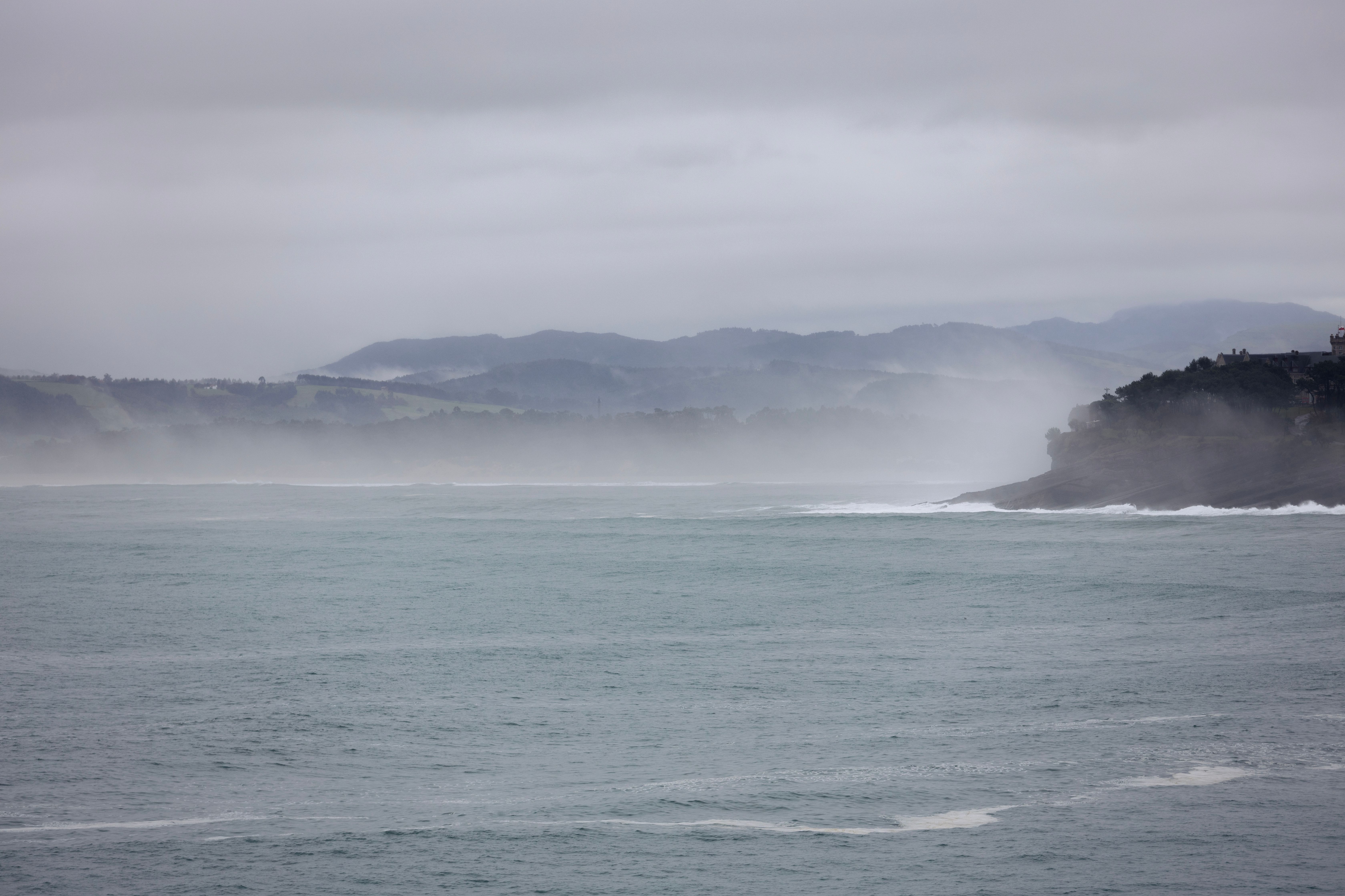Vista panorámica de un mar agitado con olas rompiendo cerca de una costa montañosa y brumosa.