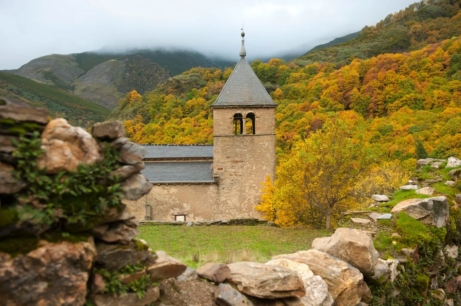 Antigua iglesia de piedra con campanario en El Bierzo, rodeada de montañas nubladas y bosques otoñales amarillos y naranjas, vista desde un muro