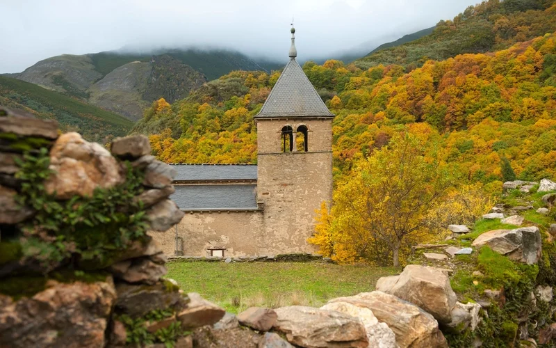 Antigua iglesia de piedra con campanario en El Bierzo, rodeada de montañas nubladas y bosques otoñales amarillos y naranjas, vista desde un muro