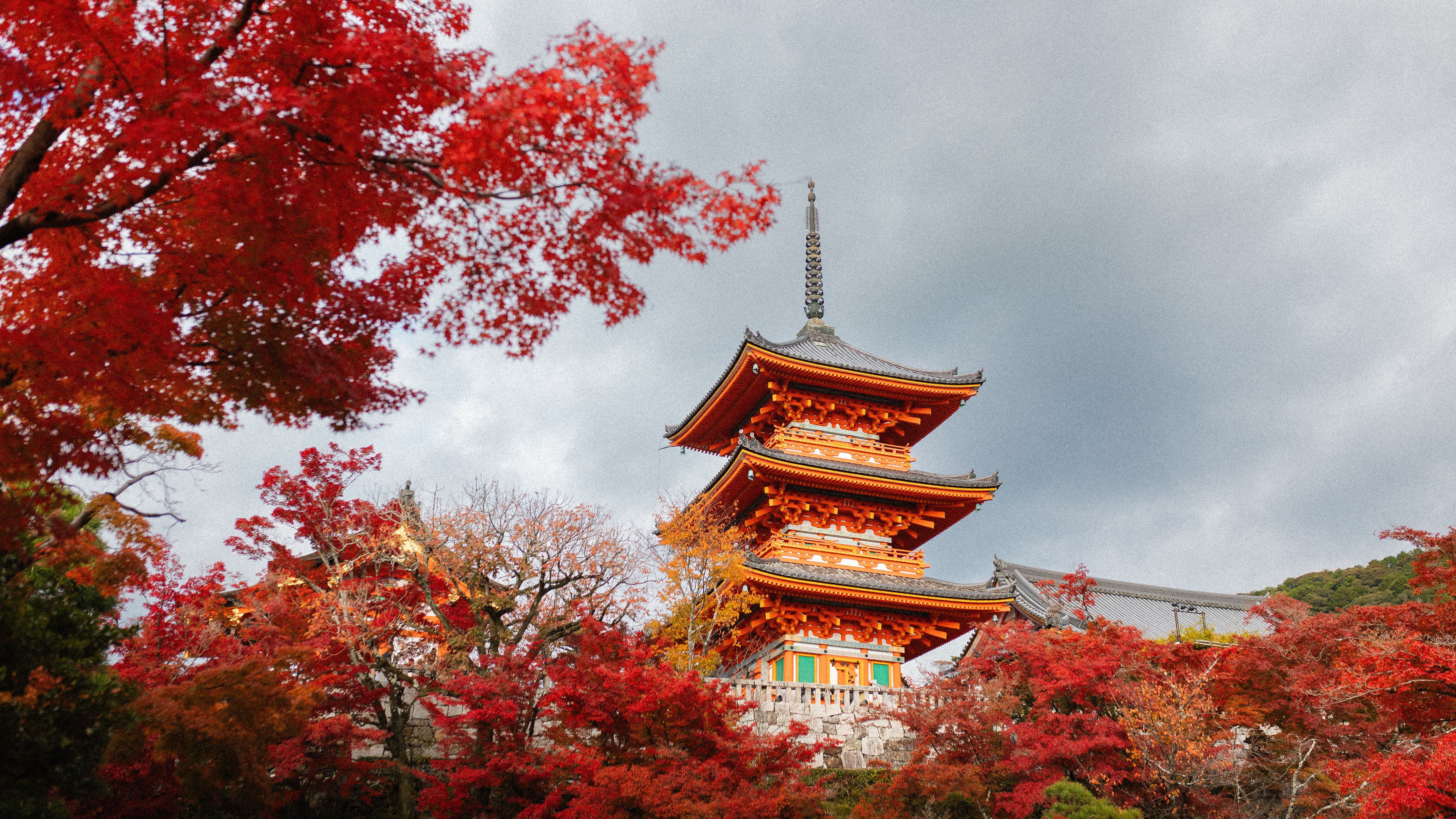 Pagoda japonesa de tres pisos en tonos naranja y rojo, rodeada de árboles con hojas otoñales.
