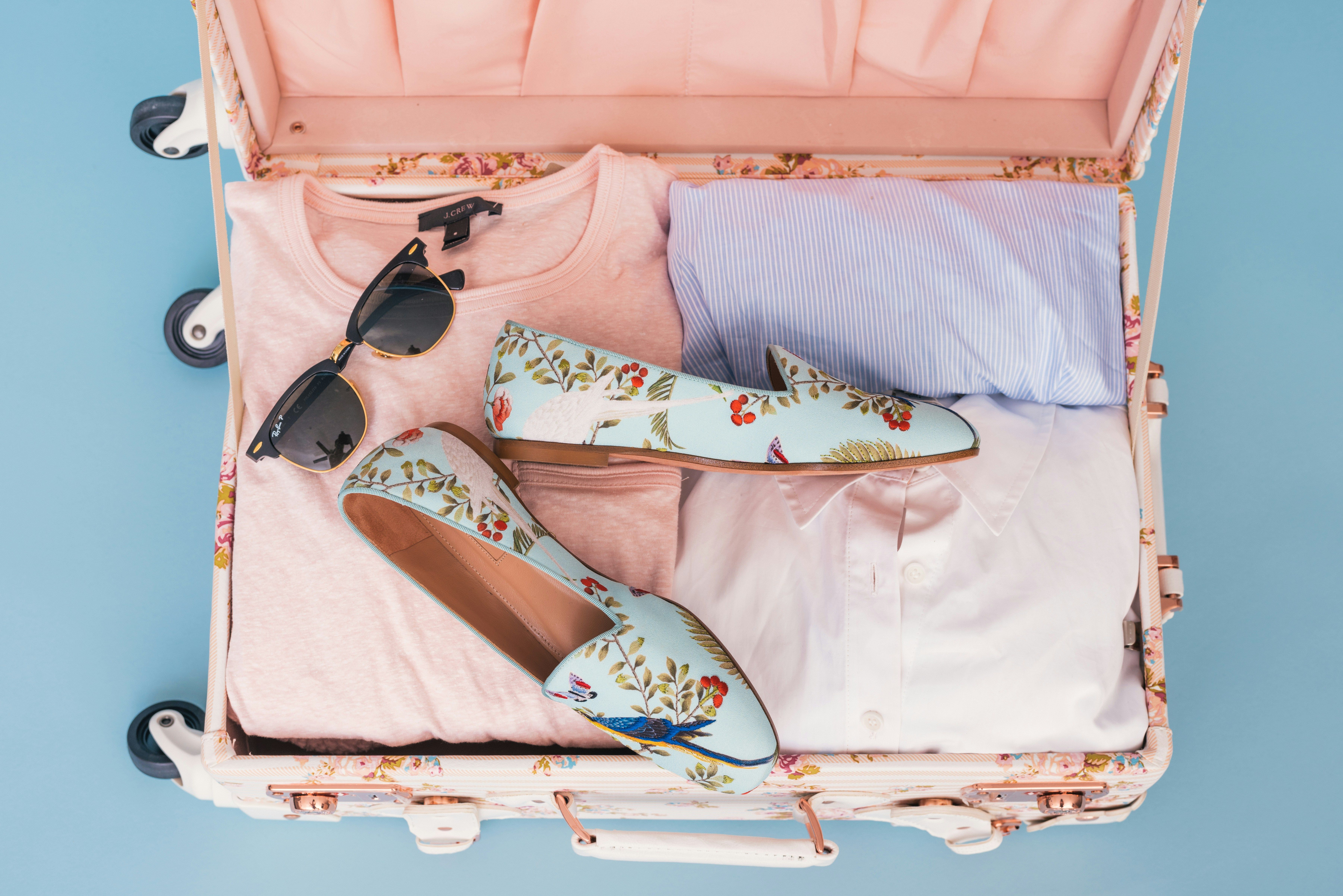 Maleta abierta con ropa doblada, un par de zapatos planos y gafas de sol sobre un fondo azul.
