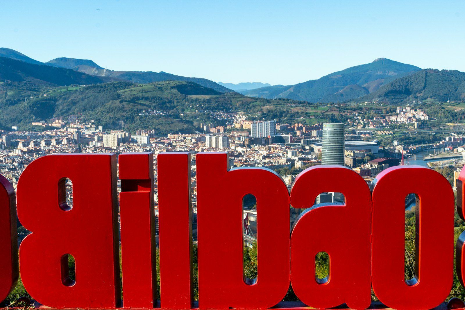 Señal roja gigante con la palabra 'Bilbao' en primer plano, vista panorámica de la ciudad, el Museo Guggenheim y montañas verdes al fondo