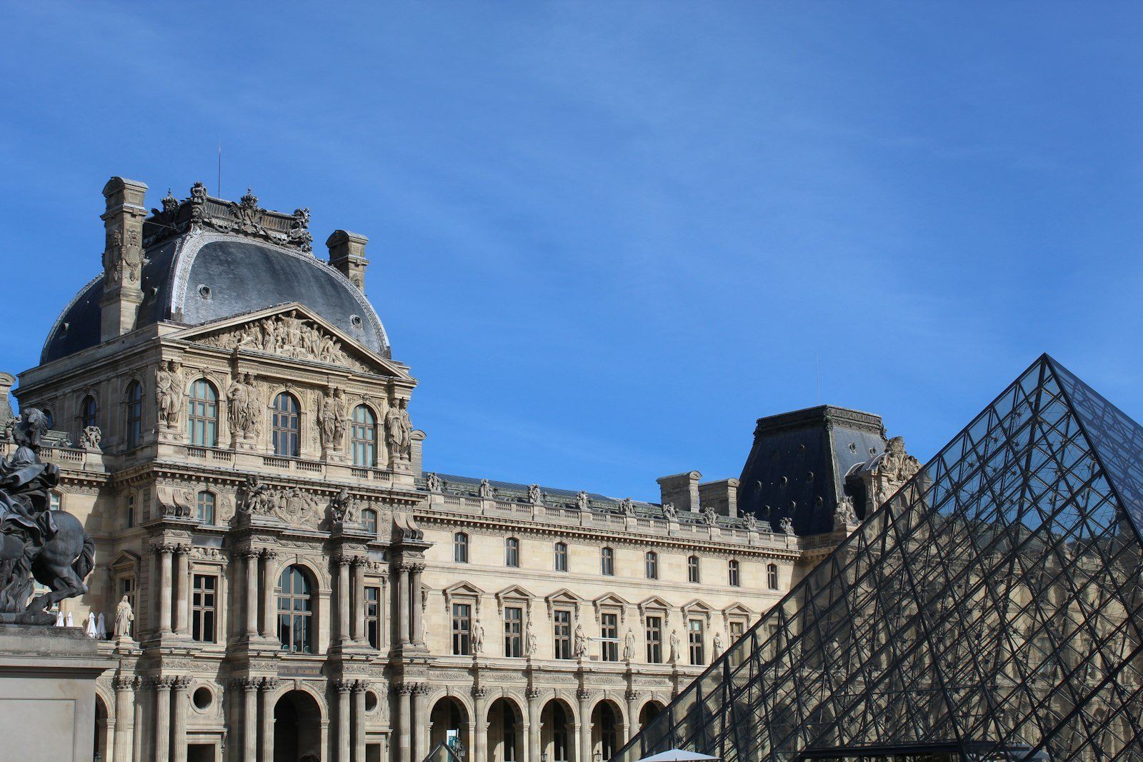 Fachada del Palacio del Louvre con la pirámide de cristal moderna al frente, bajo un cielo azul despejado