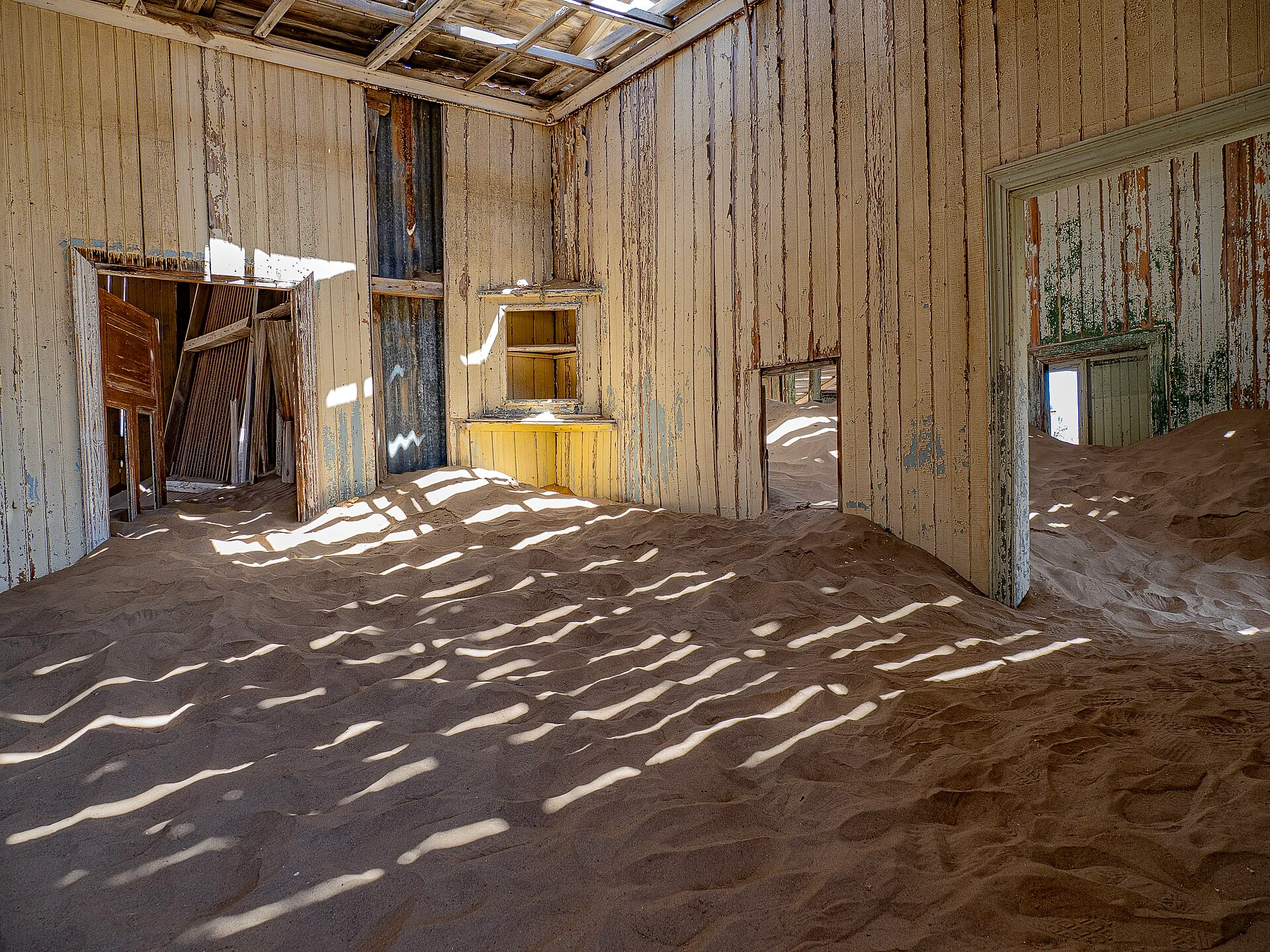 Interior de una habitación abandonada con paredes de madera descolorida y arena cubriendo el suelo.