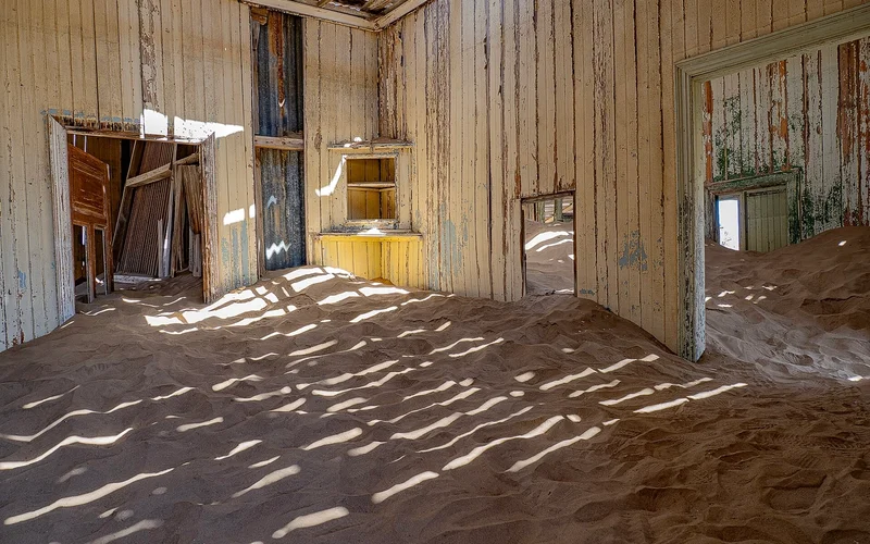 Interior de una habitación abandonada con paredes de madera descolorida y arena cubriendo el suelo.