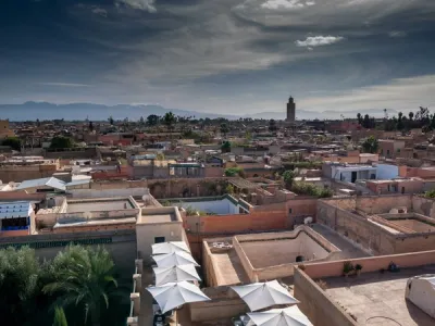 Vista panorámica de Marrakech con edificios ocre, minarete de la Koutoubia, palmeras y montañas del Atlas bajo un cielo nublado