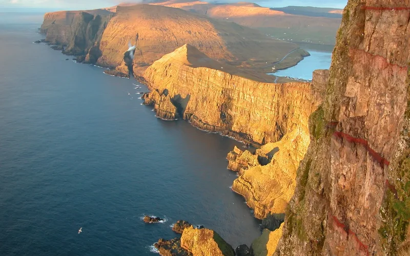 Vista panorámica de acantilados dorados en las Islas Feroe al atardecer, con mar azul y niebla envolvente