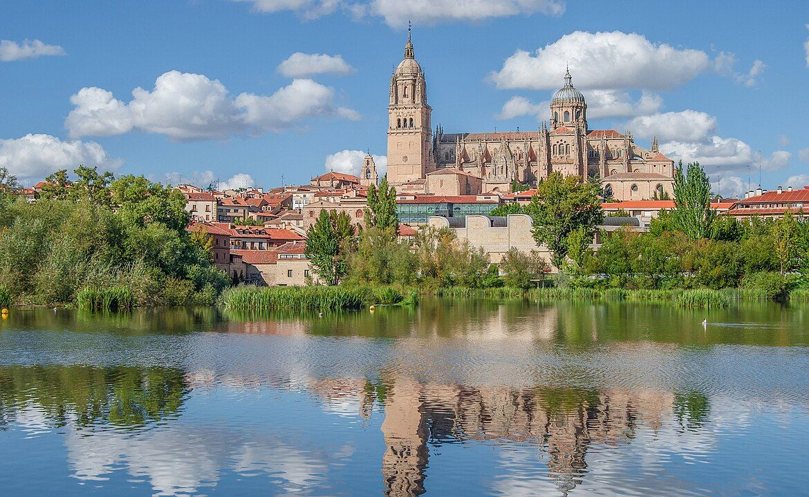 Vista de la Catedral de Salamanca y edificios históricos reflejados en el río Tormes, con vegetación ribereña y cielo azul con nubes