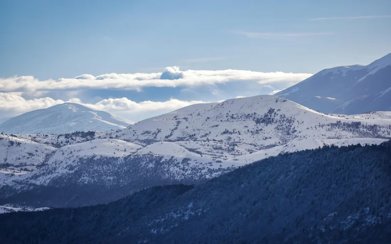 Paisaje montañoso invernal con picos nevados y bosques, bajo un cielo azul con nubes.
