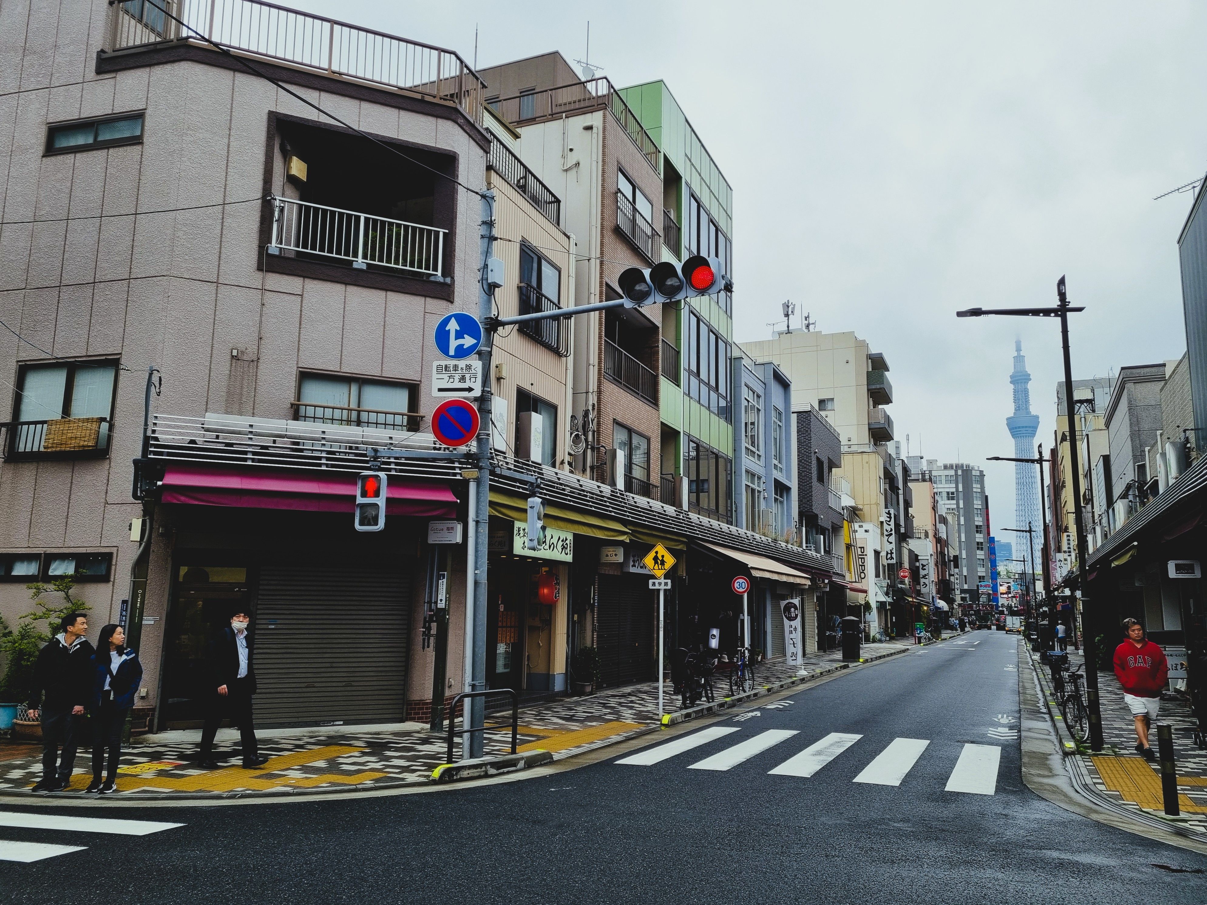 Calle urbana japonesa con edificios de varios pisos, un paso de cebra y un semáforo en rojo. Al fondo se vislumbra una torre.