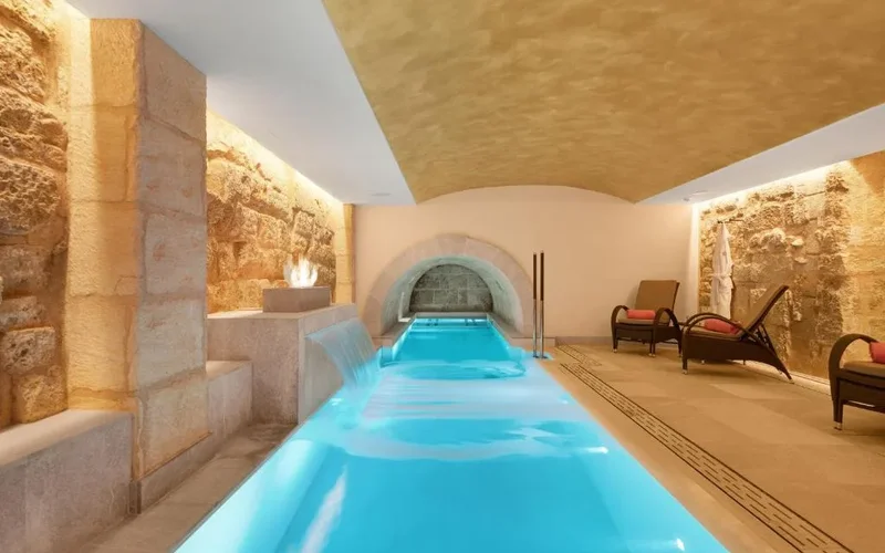Piscina interior con agua azul iluminada en un spa de piedra antigua, sillas lounge y chimenea junto a la pared