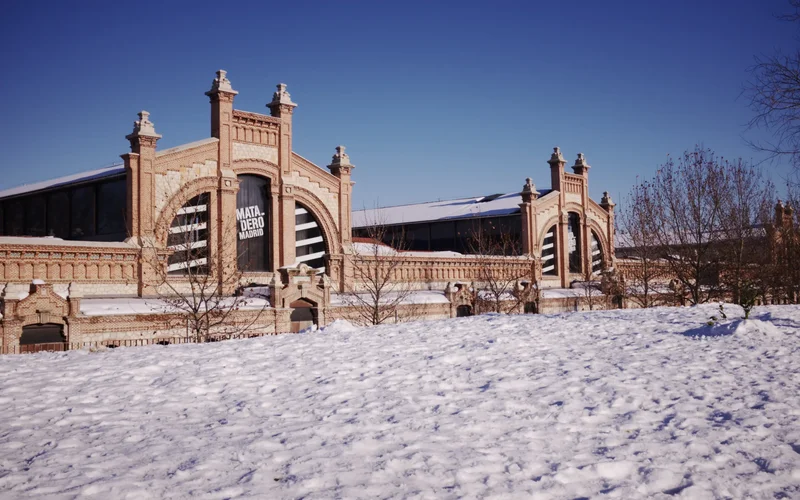 Fachada del Matadero Madrid en invierno, con edificios de ladrillo rojo, ventanas arqueadas y nieve cubriendo el suelo bajo un cielo azul