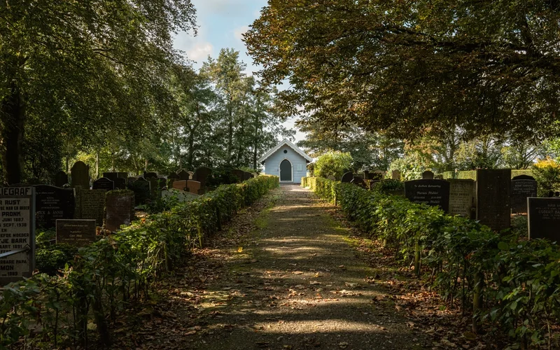Sendero arbolado con lápidas a los lados que conduce a una pequeña capilla blanca al fondo.