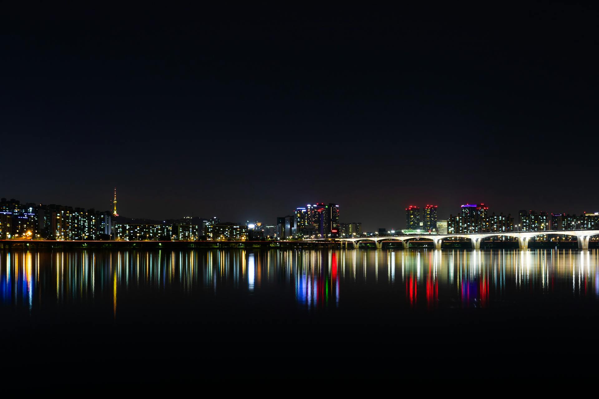 Vista nocturna del skyline de Seúl con puente iluminado y reflejos coloridos en el río