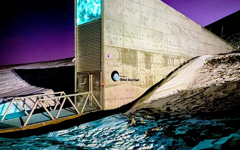 El Svalbard Global Seed Vault, estructura de concreto en colina nevada, iluminada en azul bajo cielo púrpura al atardecer