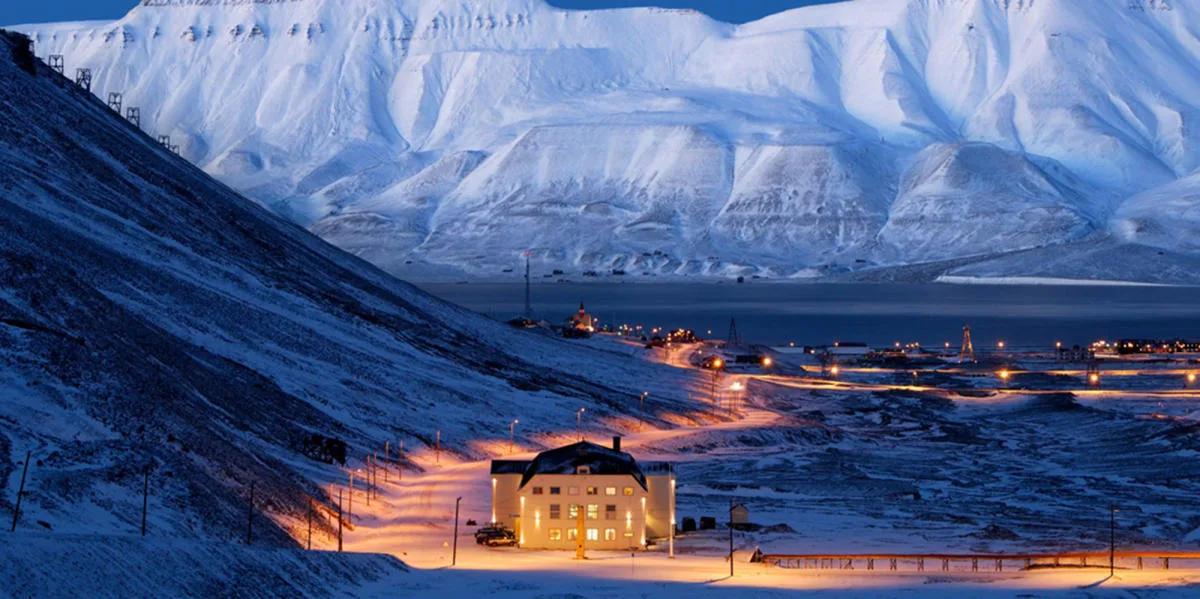 Paisaje ártico de Svalbard con montañas nevadas imponentes, un edificio iluminado en primer plano y puerto con luces en la distancia al crepúsculo