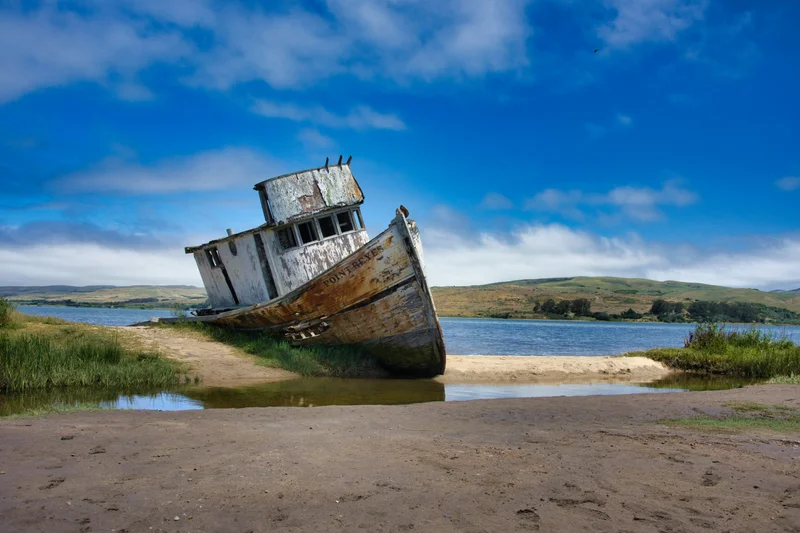 Barco de madera viejo y oxidado varado en la orilla de un lago bajo un cielo azul brillante.