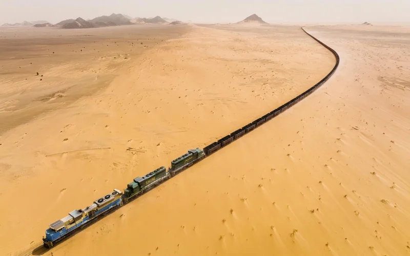 El tren de mineral serpentea por dunas doradas del Sáhara en Mauritania, con montañas difuminadas al fondo