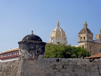 Muralla colonial de piedra en Cartagena de Indias, con torre cilíndrica negra y catedral de cúpula dorada y torres al fondo, cielo azul