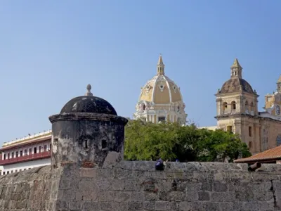 Muralla colonial de piedra en Cartagena de Indias, con torre cilíndrica negra y catedral de cúpula dorada y torres al fondo, cielo azul
