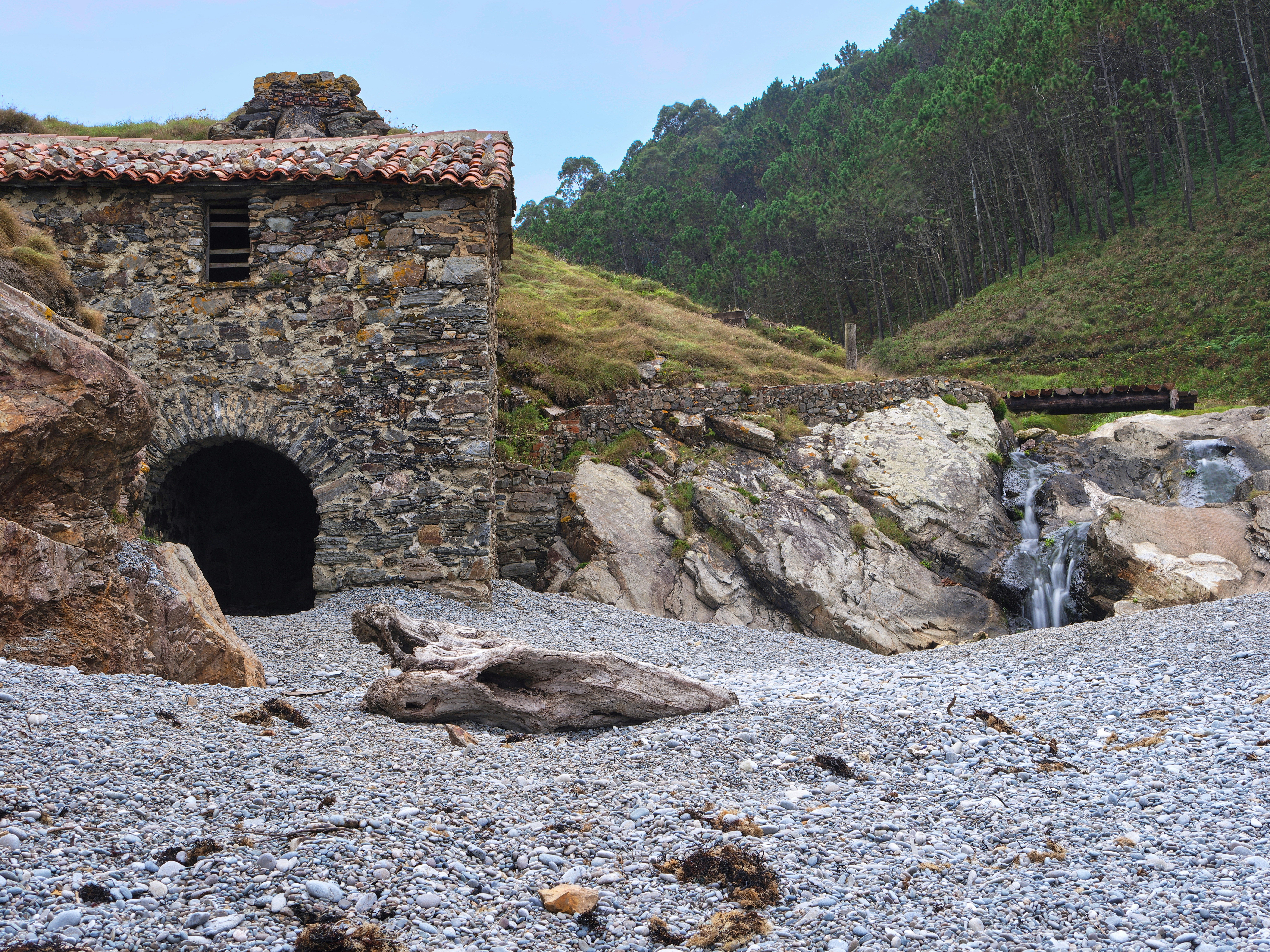 Edificio de piedra con techo de tejas junto a una cascada pequeña y un lecho de río pedregoso.