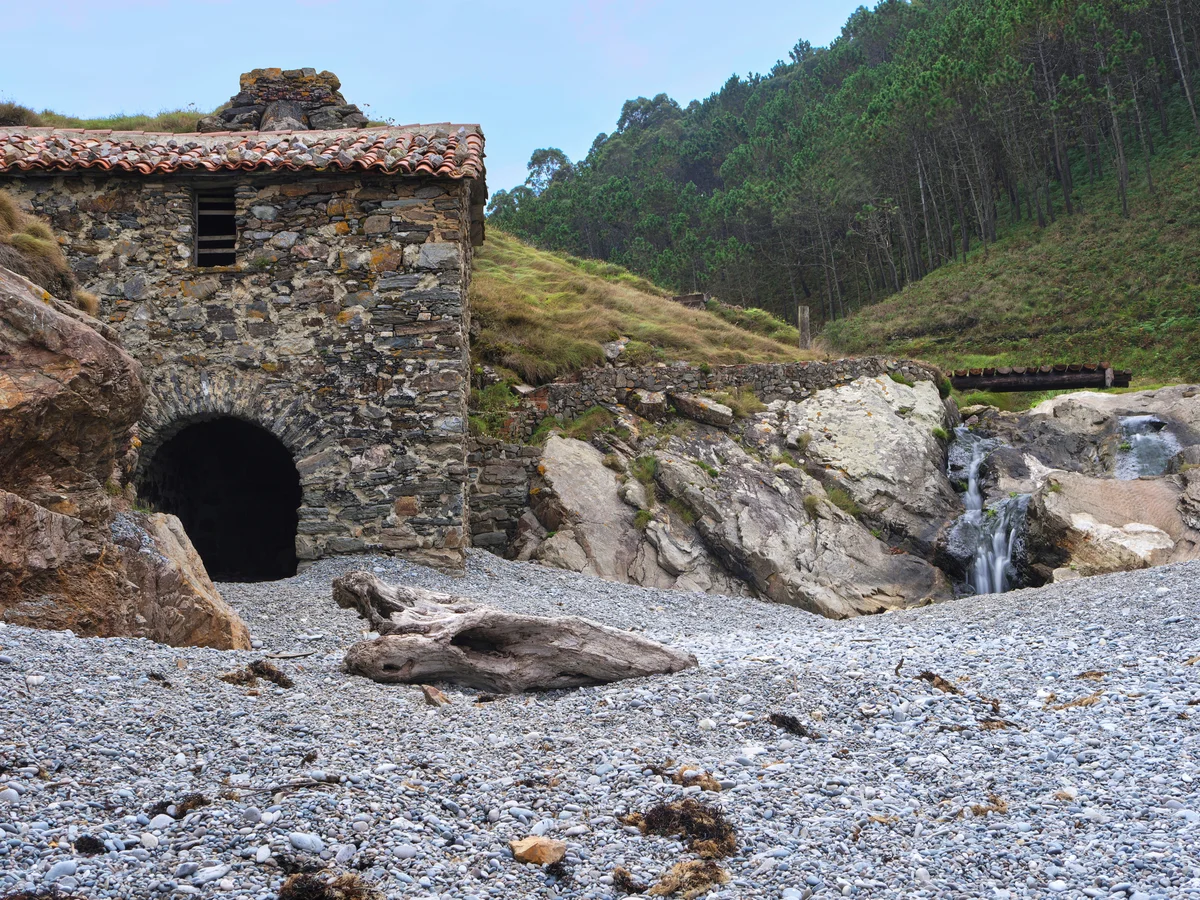 Edificio de piedra con techo de tejas junto a una cascada pequeña y un lecho de río pedregoso.