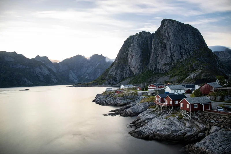 Casas rojas y blancas sobre rocas junto a un fiordo con montañas al fondo bajo un cielo nublado.