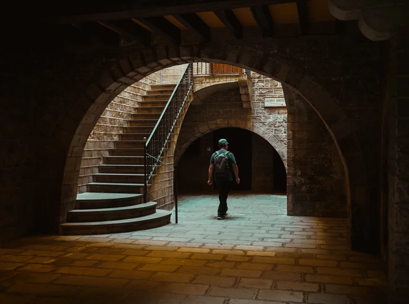 Persona caminando por un pasillo abovedado de piedra con una escalera curva a la izquierda.