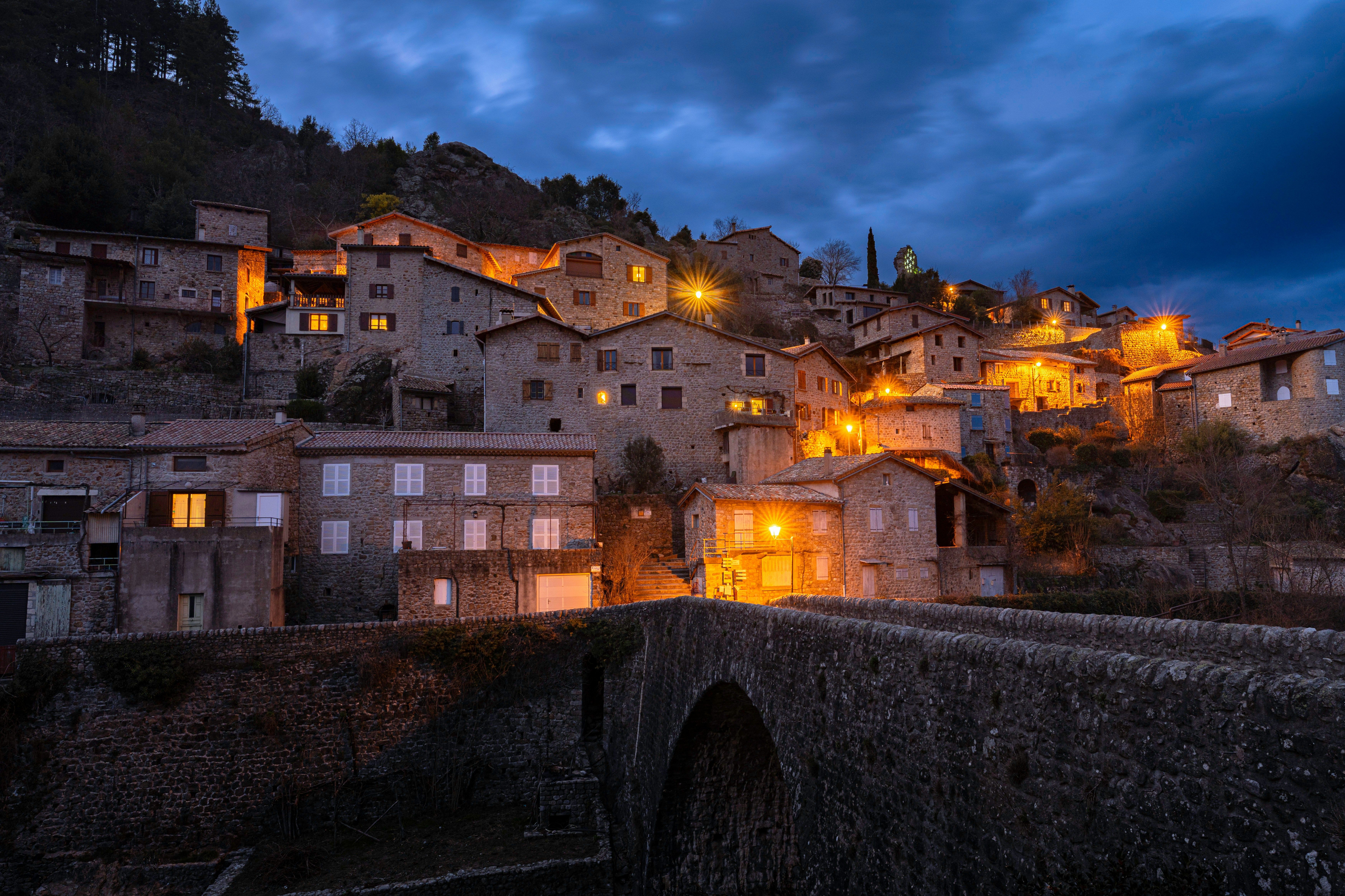 Vista nocturna de un pueblo de piedra en la ladera de una montaña, con luces encendidas en las ventanas y un puente de piedra en primer plano.