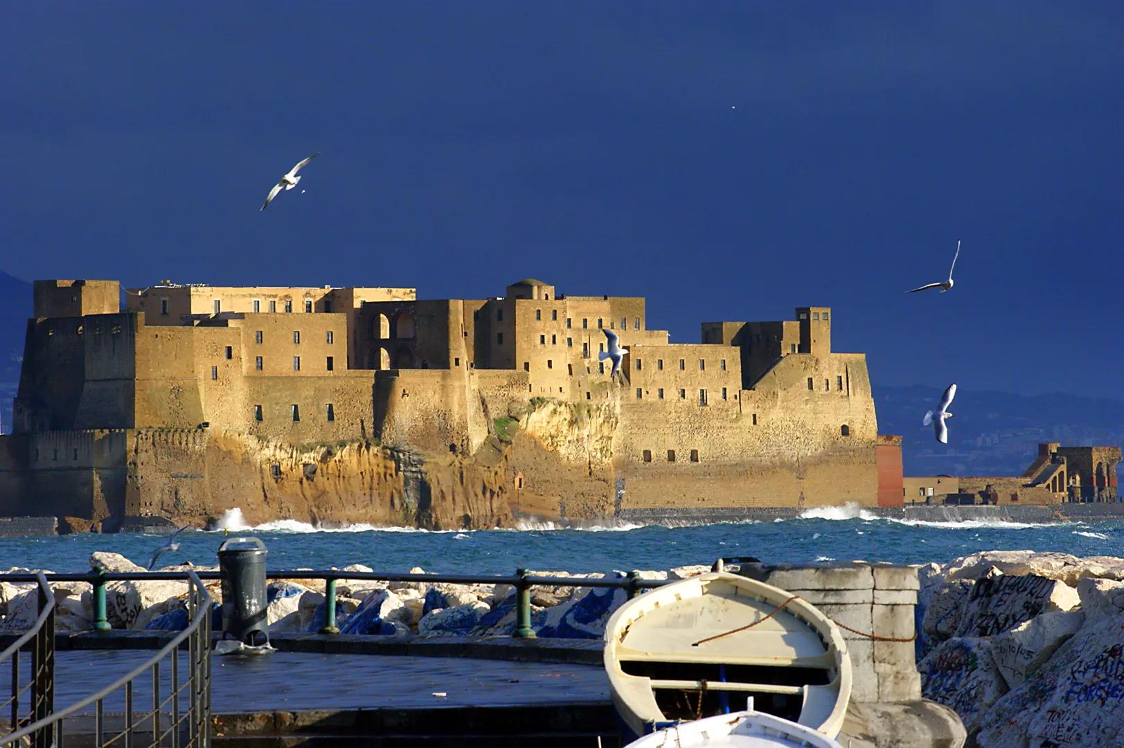 Castel dell'Ovo en Nápoles al atardecer, con murallas doradas sobre el mar, gaviotas volando y un bote en el muelle