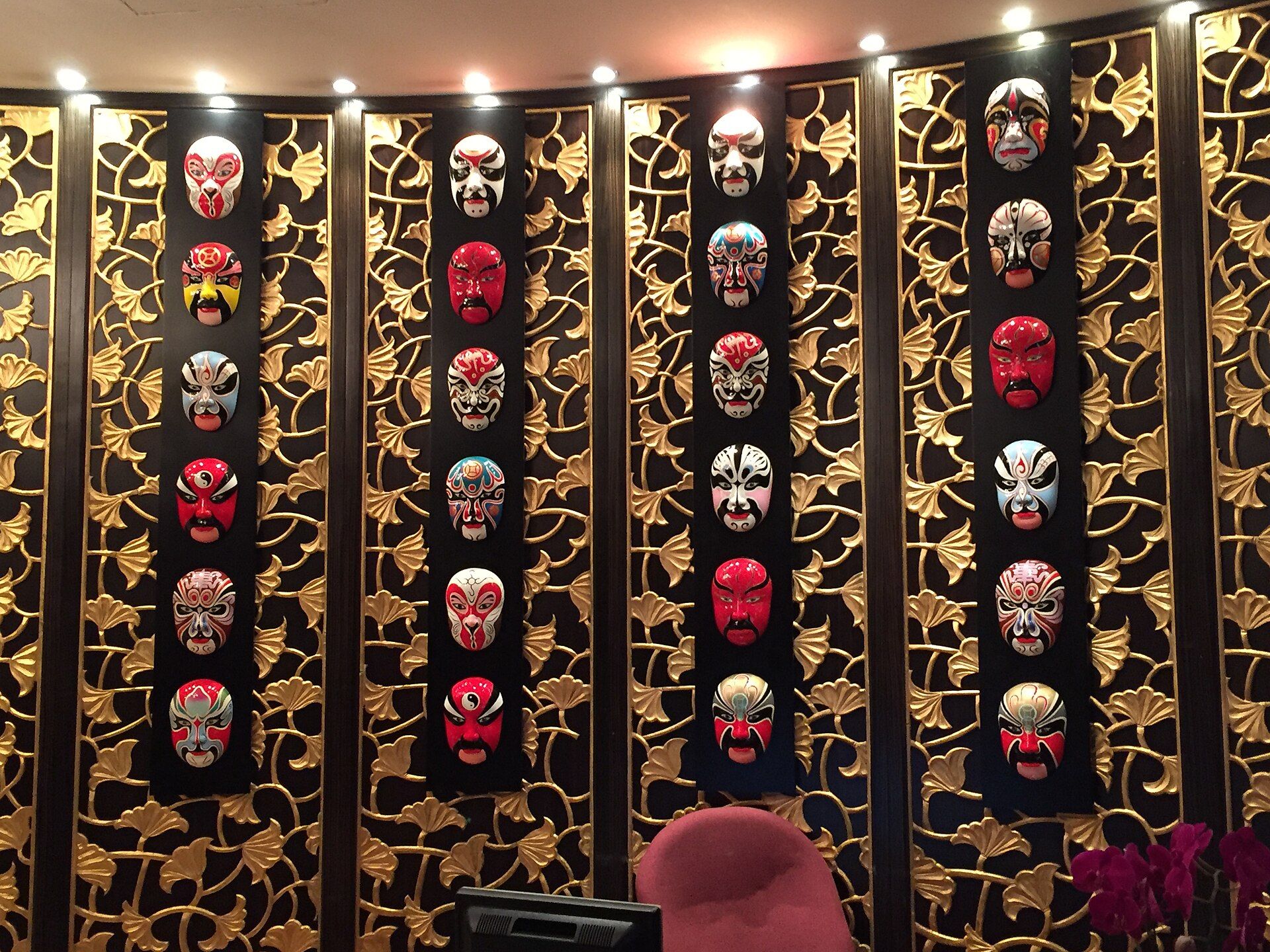 Mural decorativo con múltiples máscaras de ópera chinas de colores vibrantes sobre un fondo oscuro y dorado.