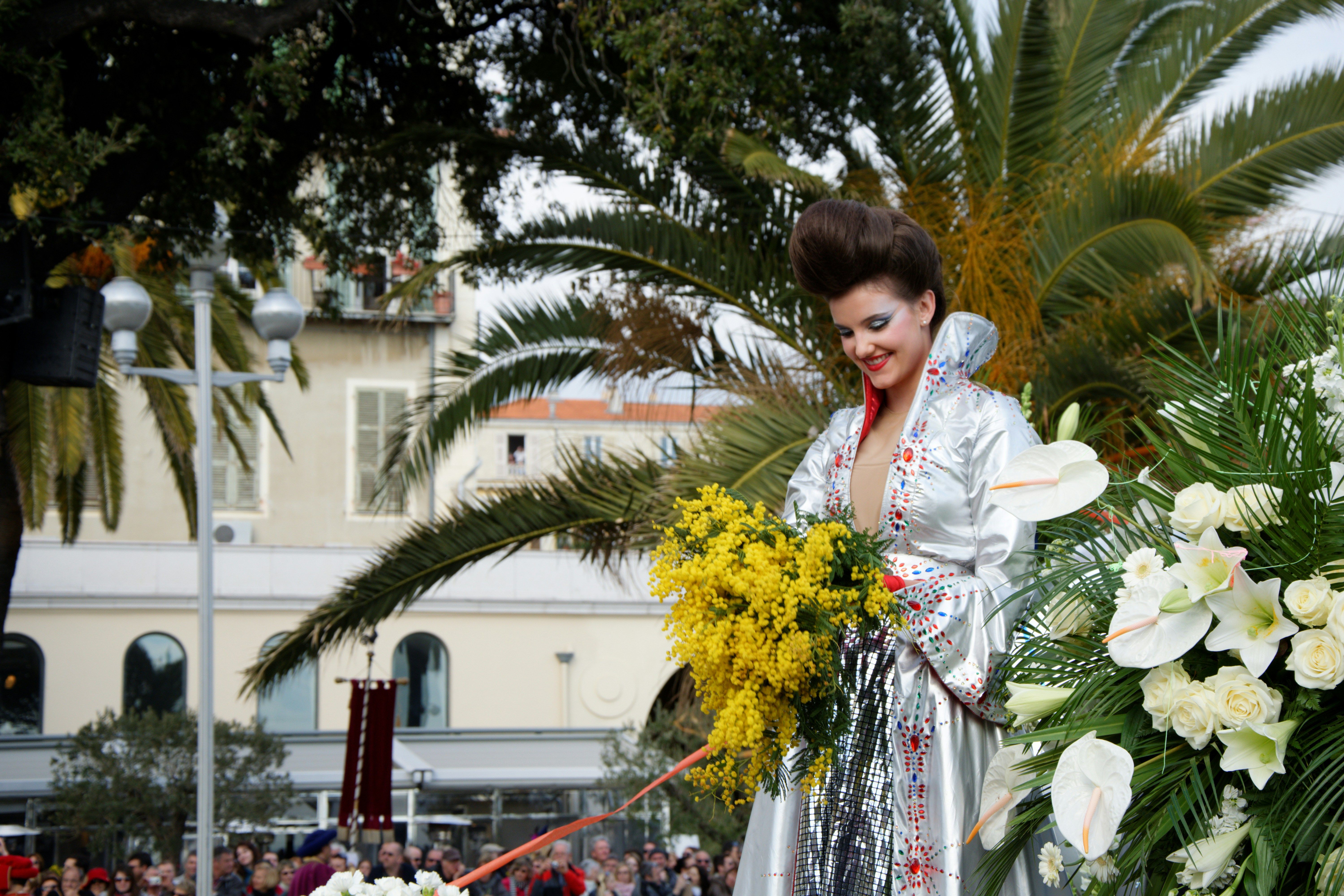 Mujer con peinado alto y vestido plateado brillante sosteniendo flores amarillas en un desfile floral.