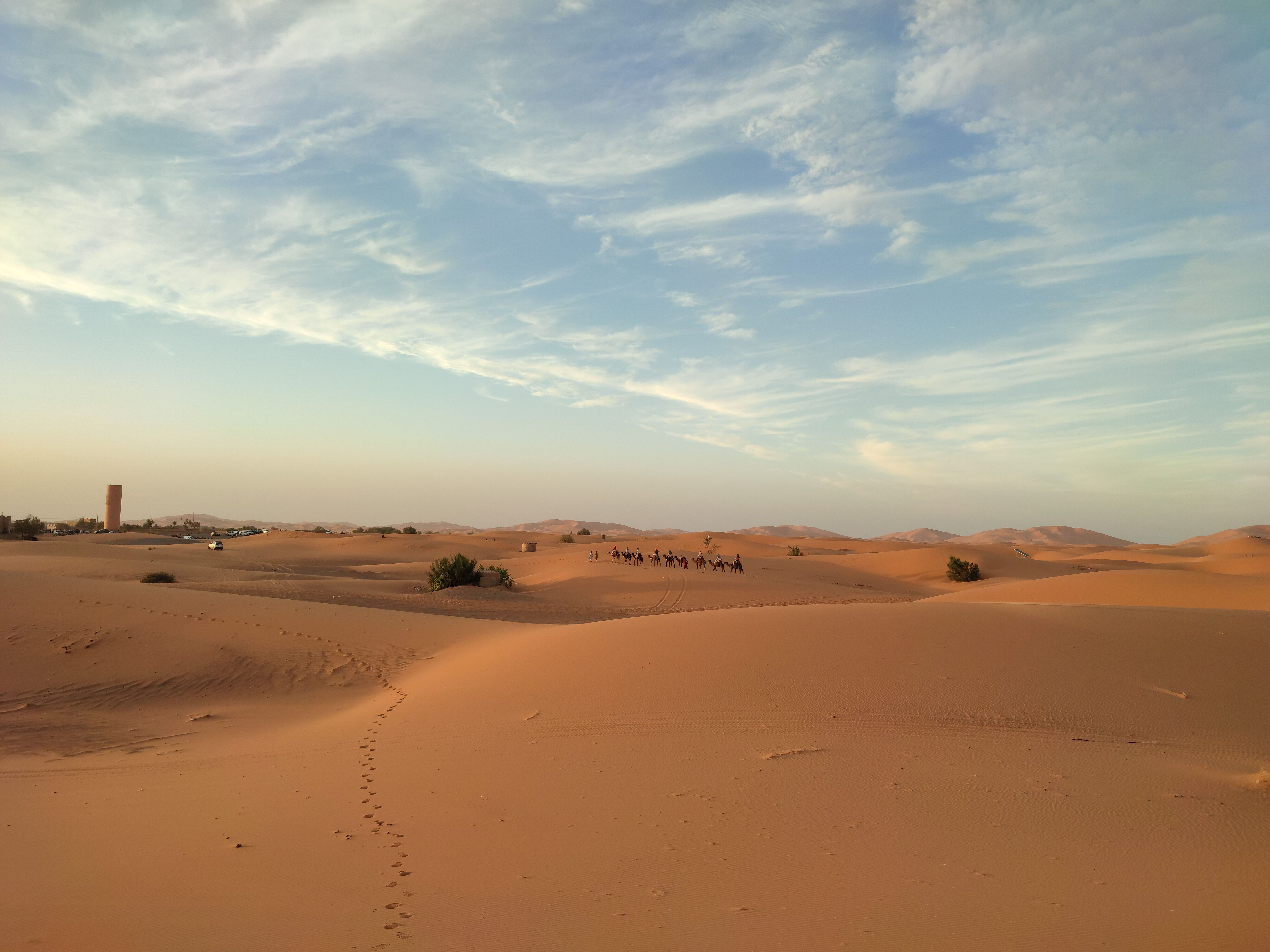 Amplio paisaje desértico con dunas de arena, un cielo azul con nubes y una caravana de camellos a lo lejos.