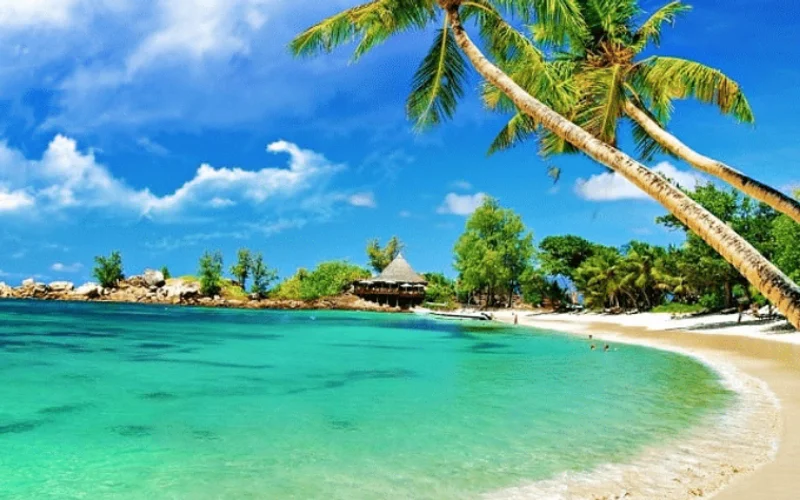 Playa tropical de arena blanca con aguas turquesas, palmeras inclinadas y cielo azul nublado en Sunset Town, Phu Quoc