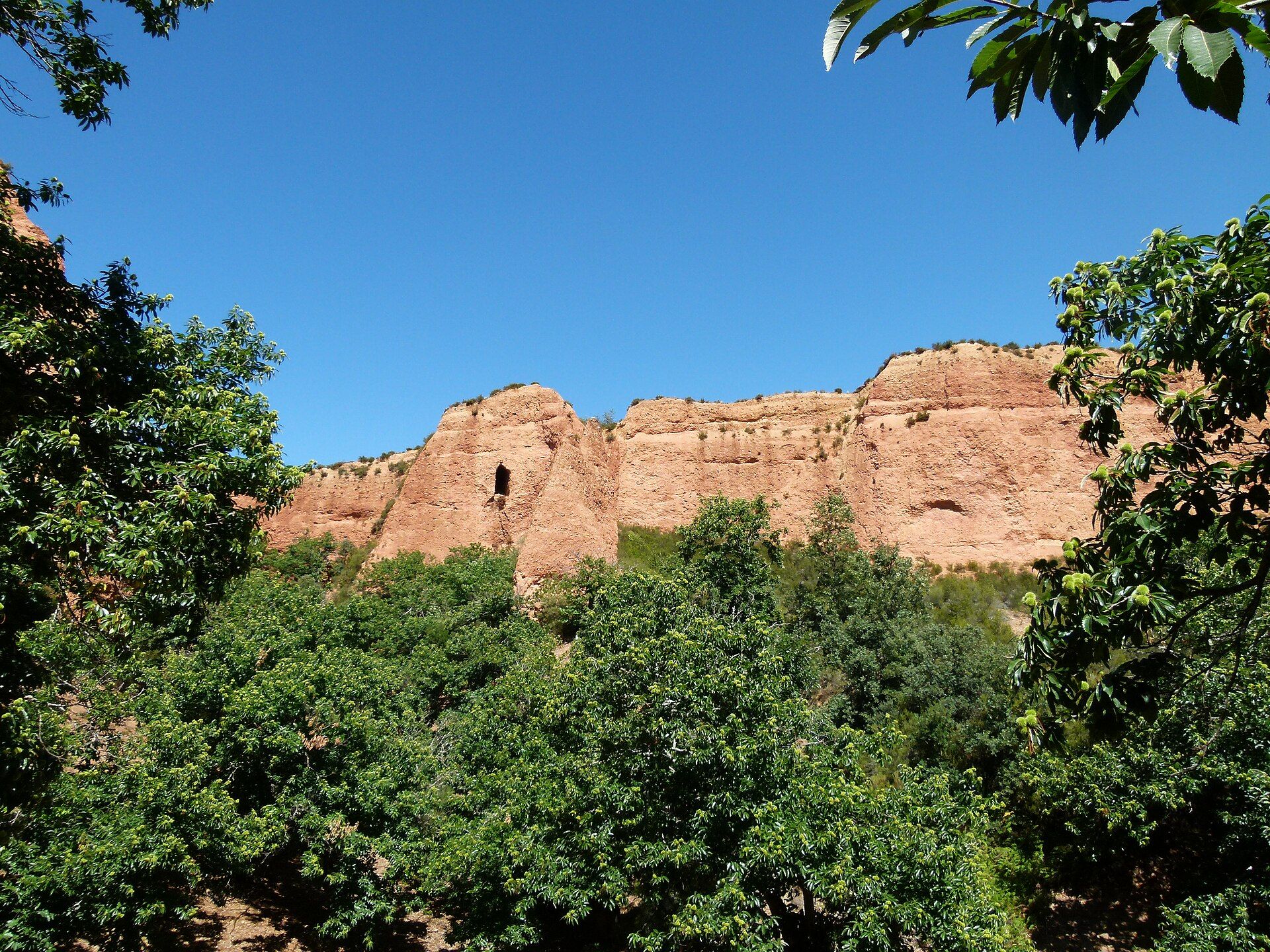 Formaciones rocosas rojizas de Las Médulas con vegetación verde y cielo azul claro.