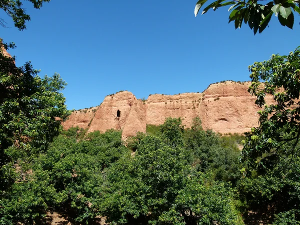 Formaciones rocosas rojizas de Las Médulas con vegetación verde y cielo azul claro.