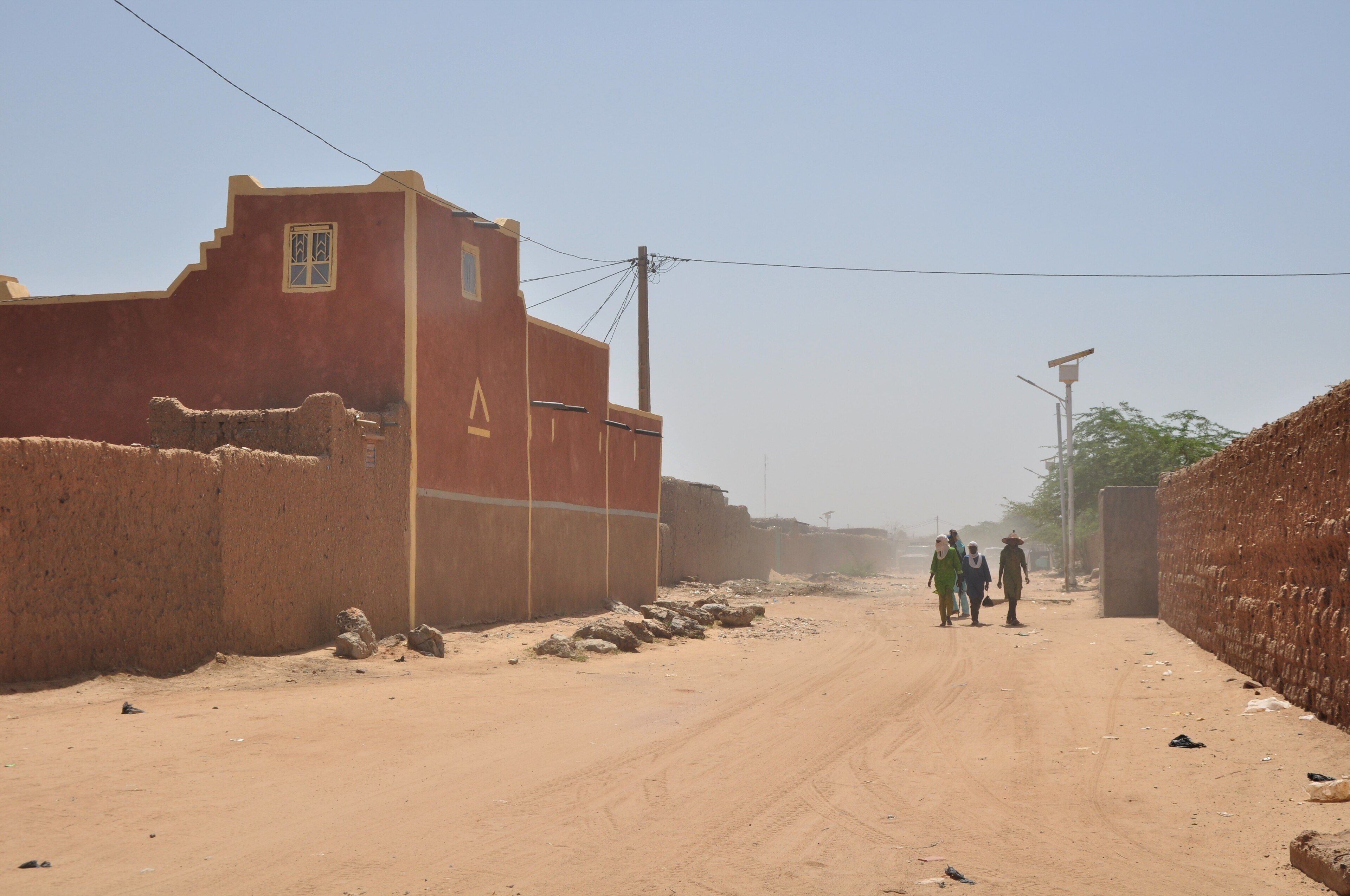 Calle de arena entre muros de adobe y una casa de fachada roja y ocre, con tres personas caminando al fondo bajo un cielo con calima.