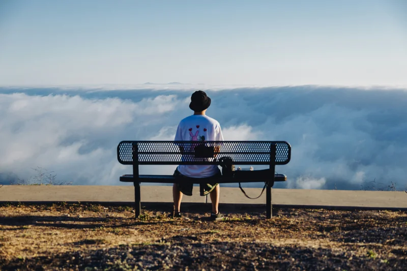 Una persona sentada de espaldas en un banco de metal, mirando un mar de nubes.