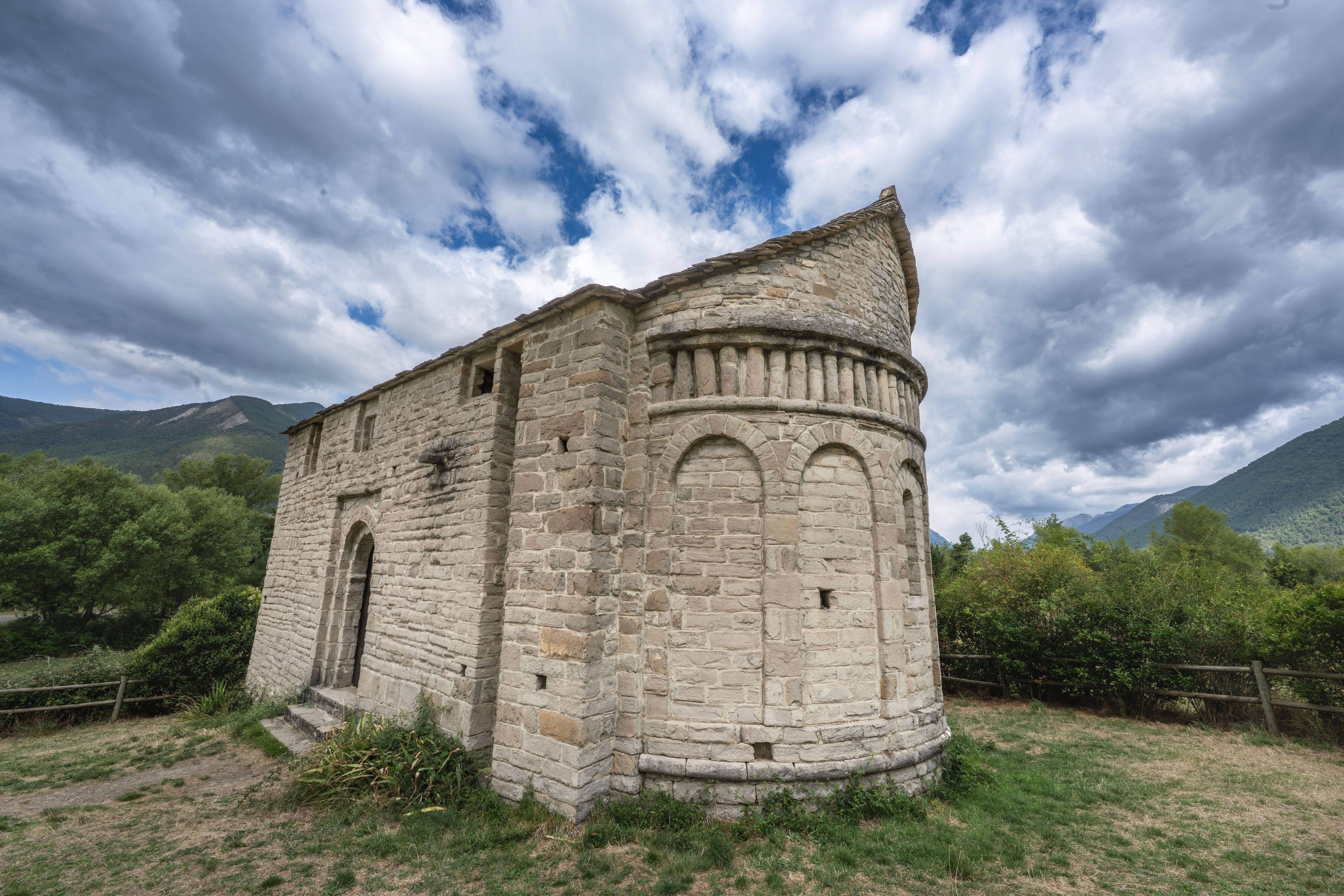Iglesia románica de piedra con ábside semicircular y montañas verdes bajo un cielo nublado.