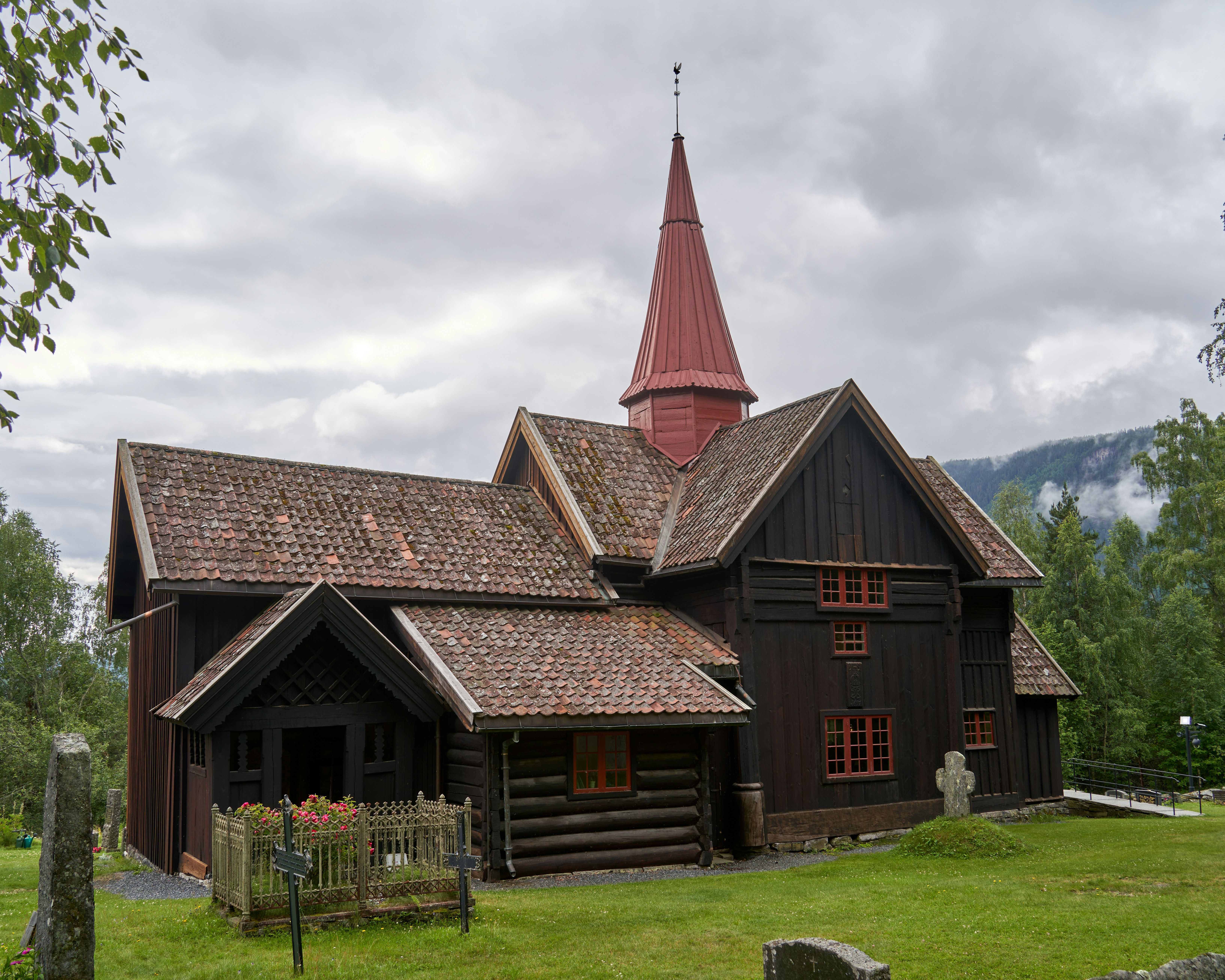 Antigua iglesia de madera oscura con tejado de tejas y aguja roja, rodeada de césped y árboles.