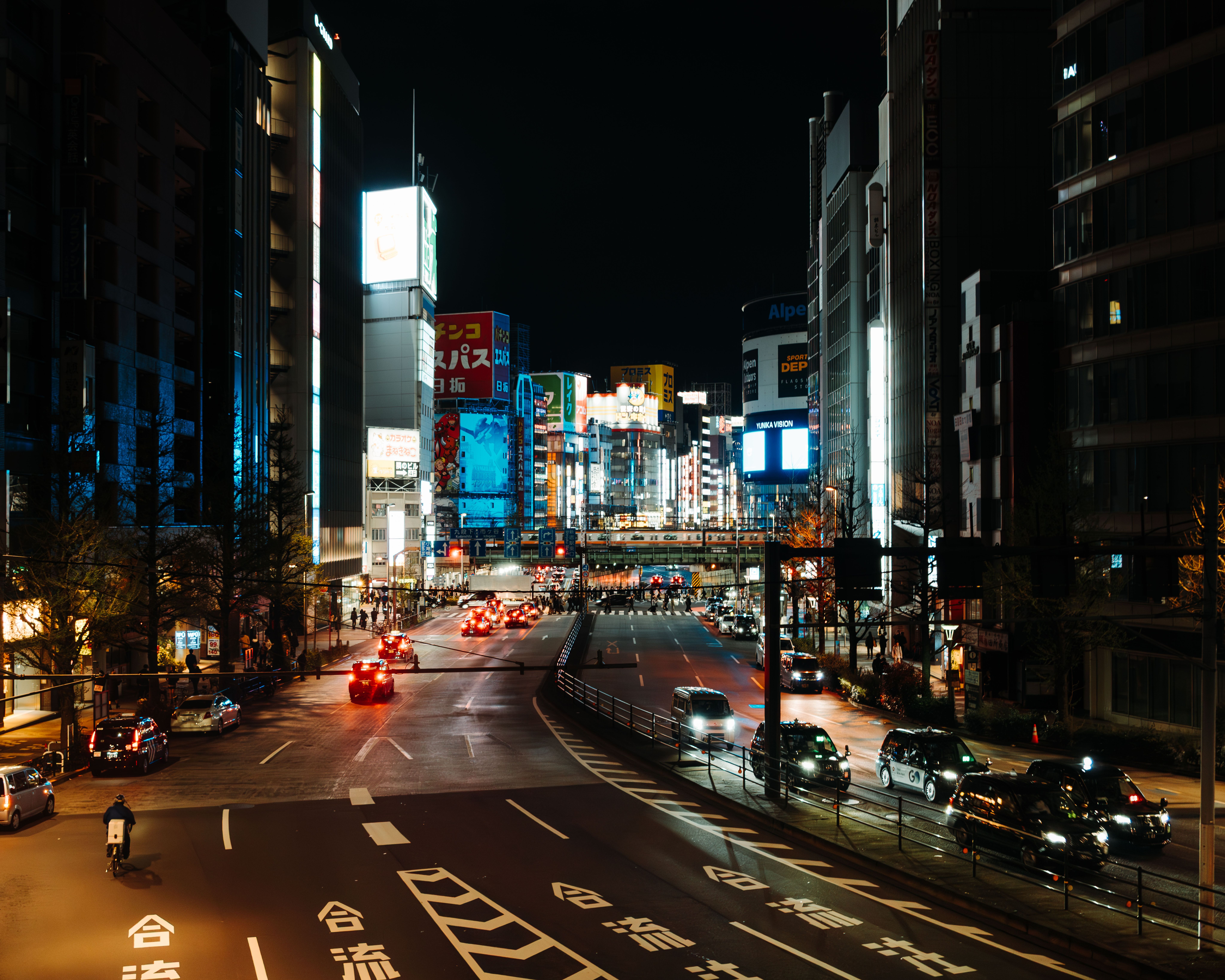 Vista nocturna de una calle con tráfico y edificios iluminados por letreros de neón en una ciudad japonesa.