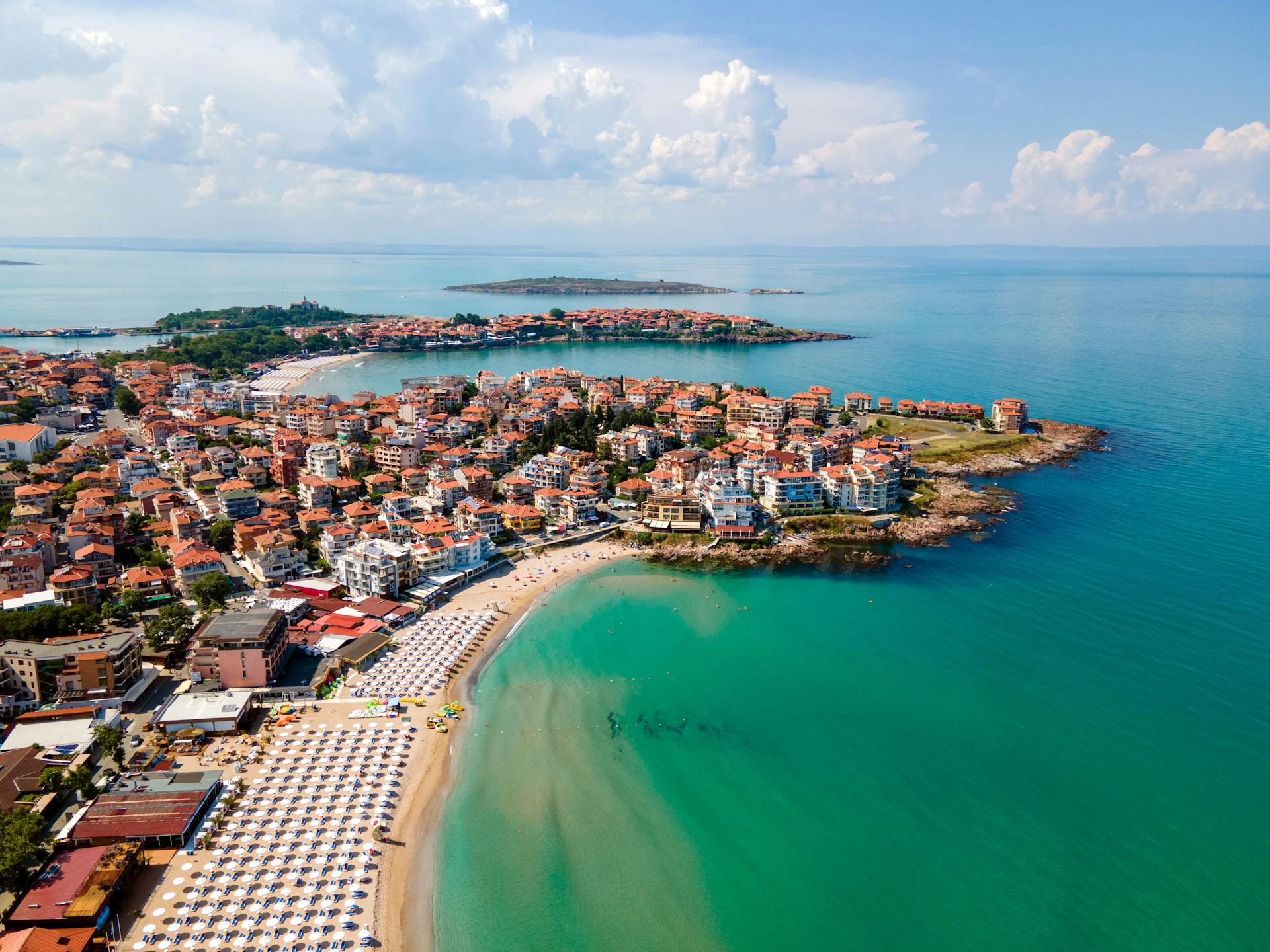 Vista aérea de Burgas con playa de arena, sombrillas blancas, edificios rojos y mar turquesa bajo nubes
