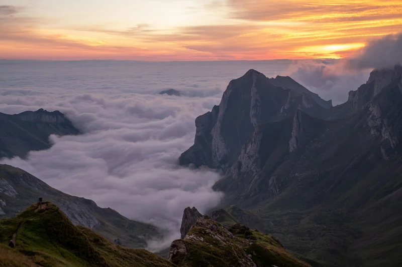Montañas escarpadas emergiendo de un mar de nubes al atardecer con un cielo anaranjado.
