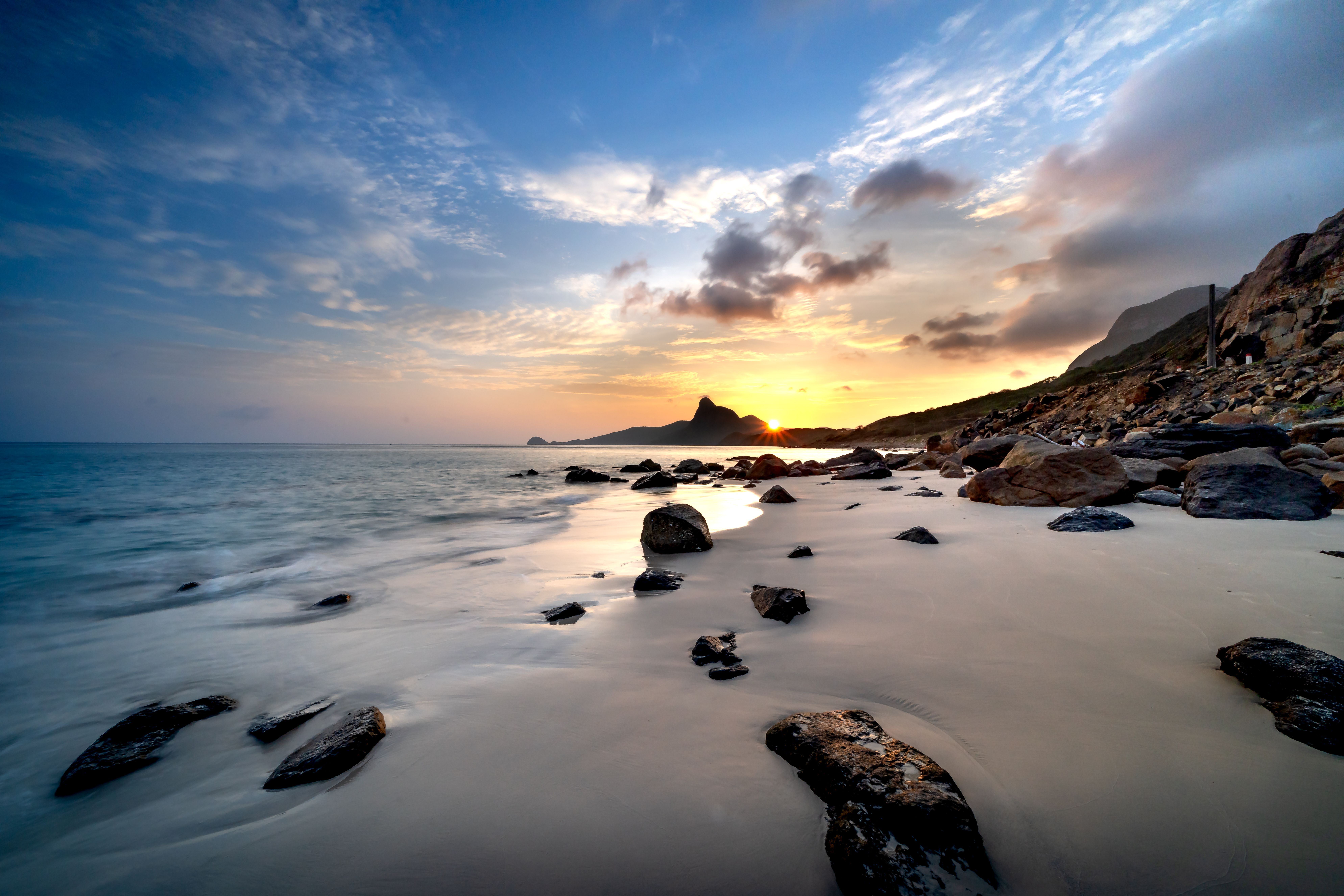 Playa rocosa al atardecer con el sol poniéndose sobre el mar y montañas al fondo.