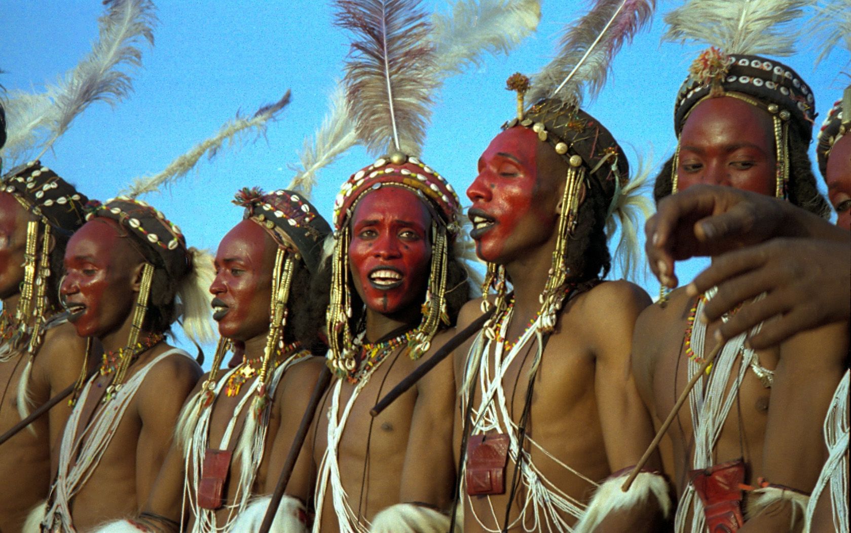Primer plano de jóvenes con la cara pintada de rojo, labios negros, tocados con conchas y plumas blancas, cantando con los dientes a la vista.