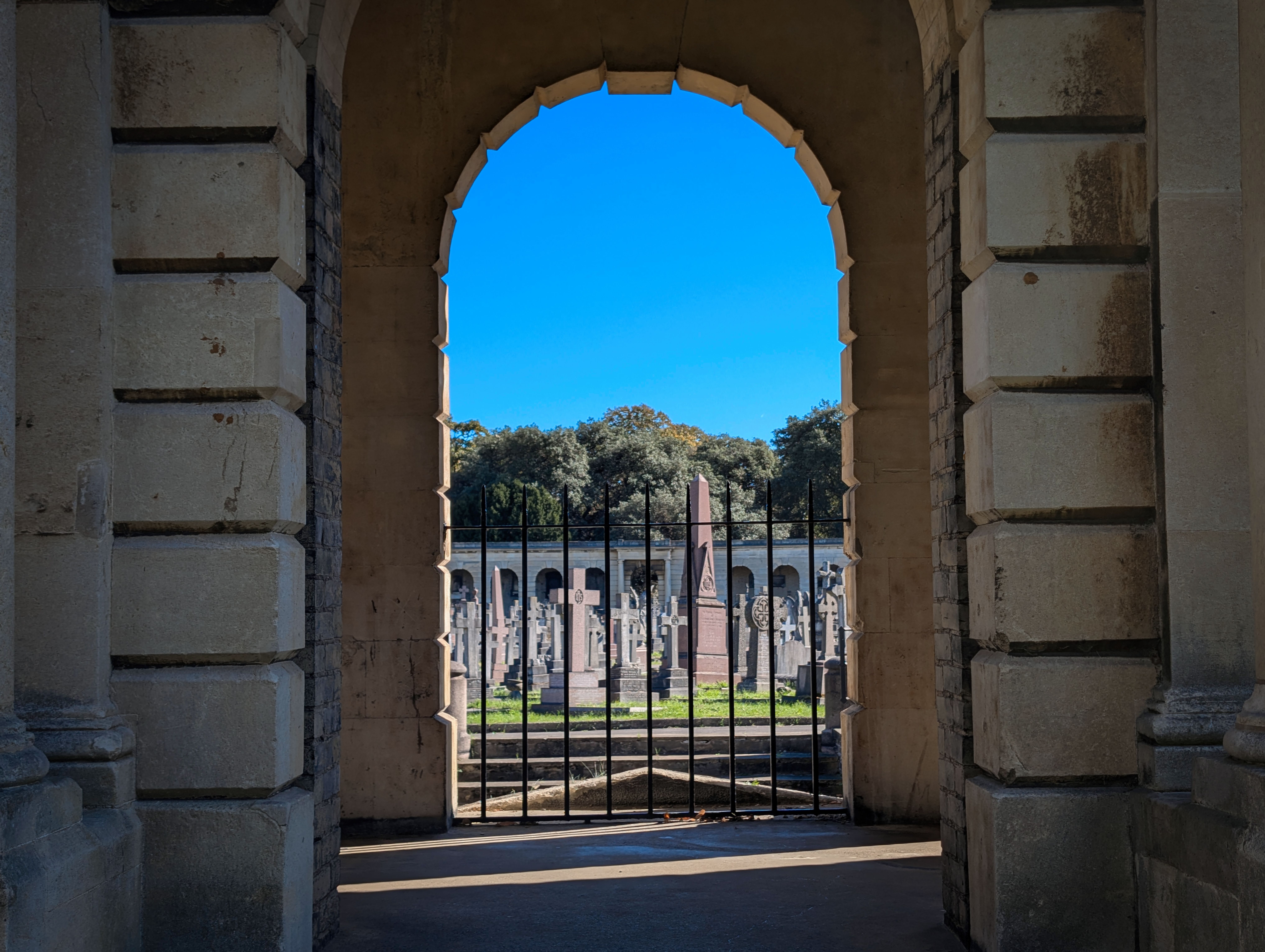 Vista a través de un arco de piedra hacia un cementerio con lápidas, cruces y árboles.